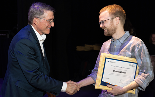 Two people shaking hands and smile at one another as the one on the right holds a plaque.