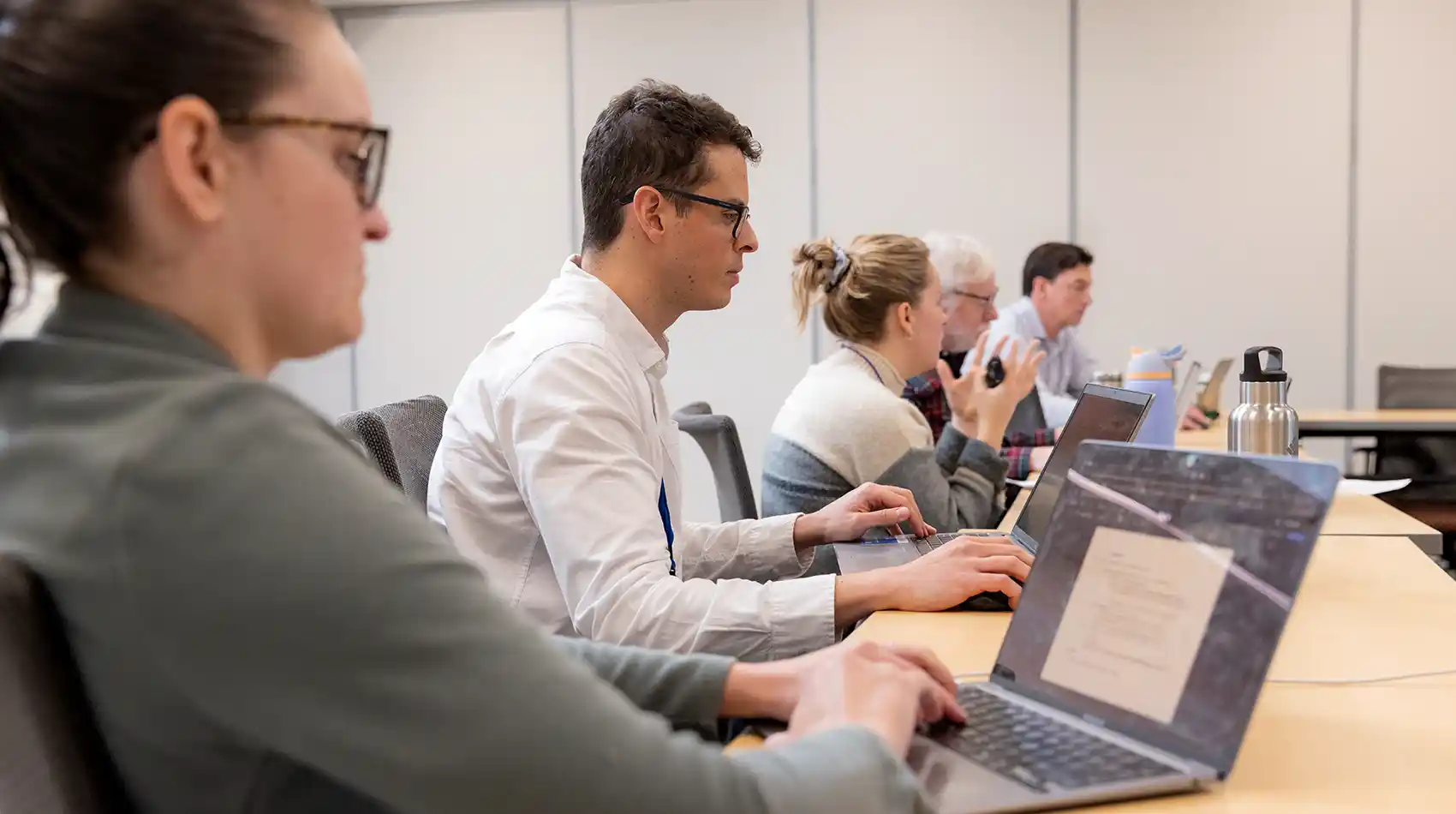 Several people sit at a desk with their laptops open.