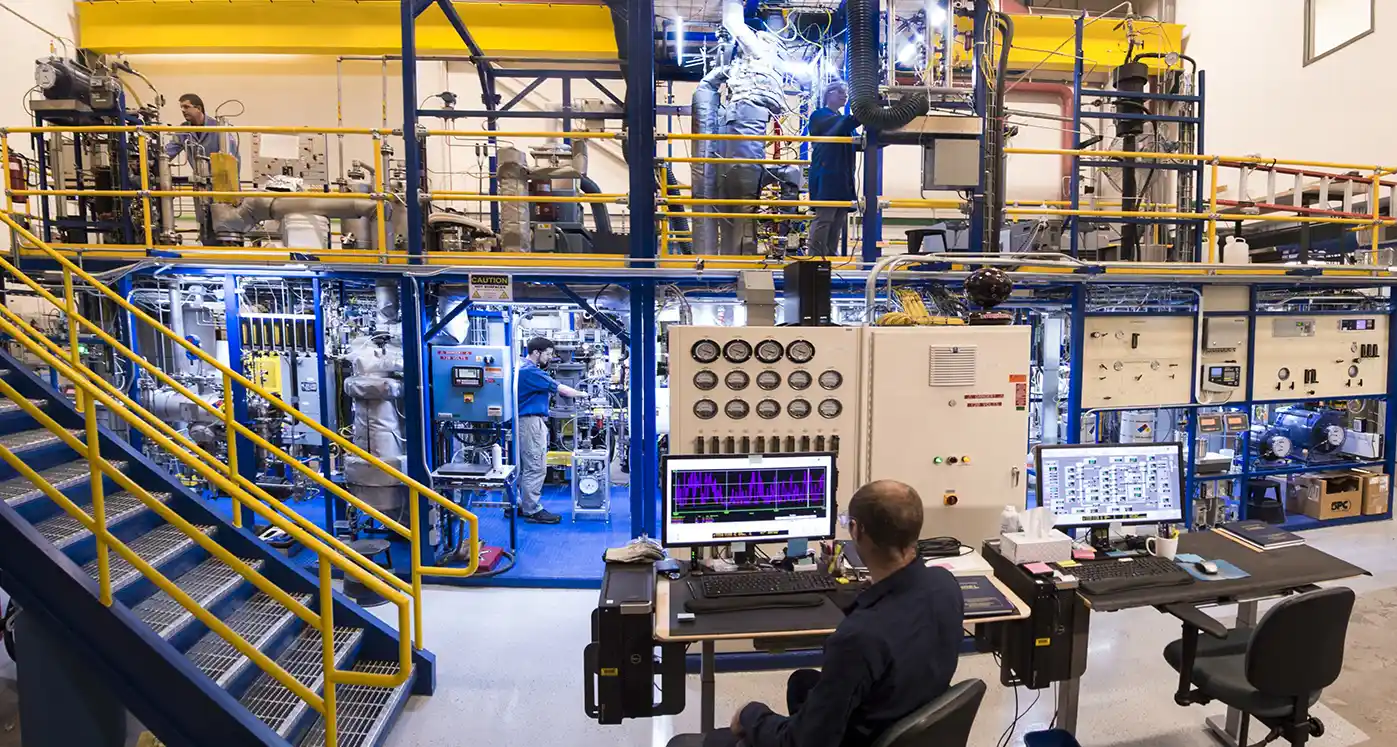 Several engineers work on a large, multilevel Davison circulating riser, with laptops on tall tables in the center. The riser features colorful scaffolding, pipes, tubes, and control panels with dials and monitors.