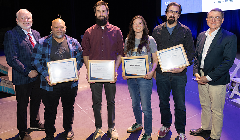 Four people pose with plaques as two people stand to their left and right.