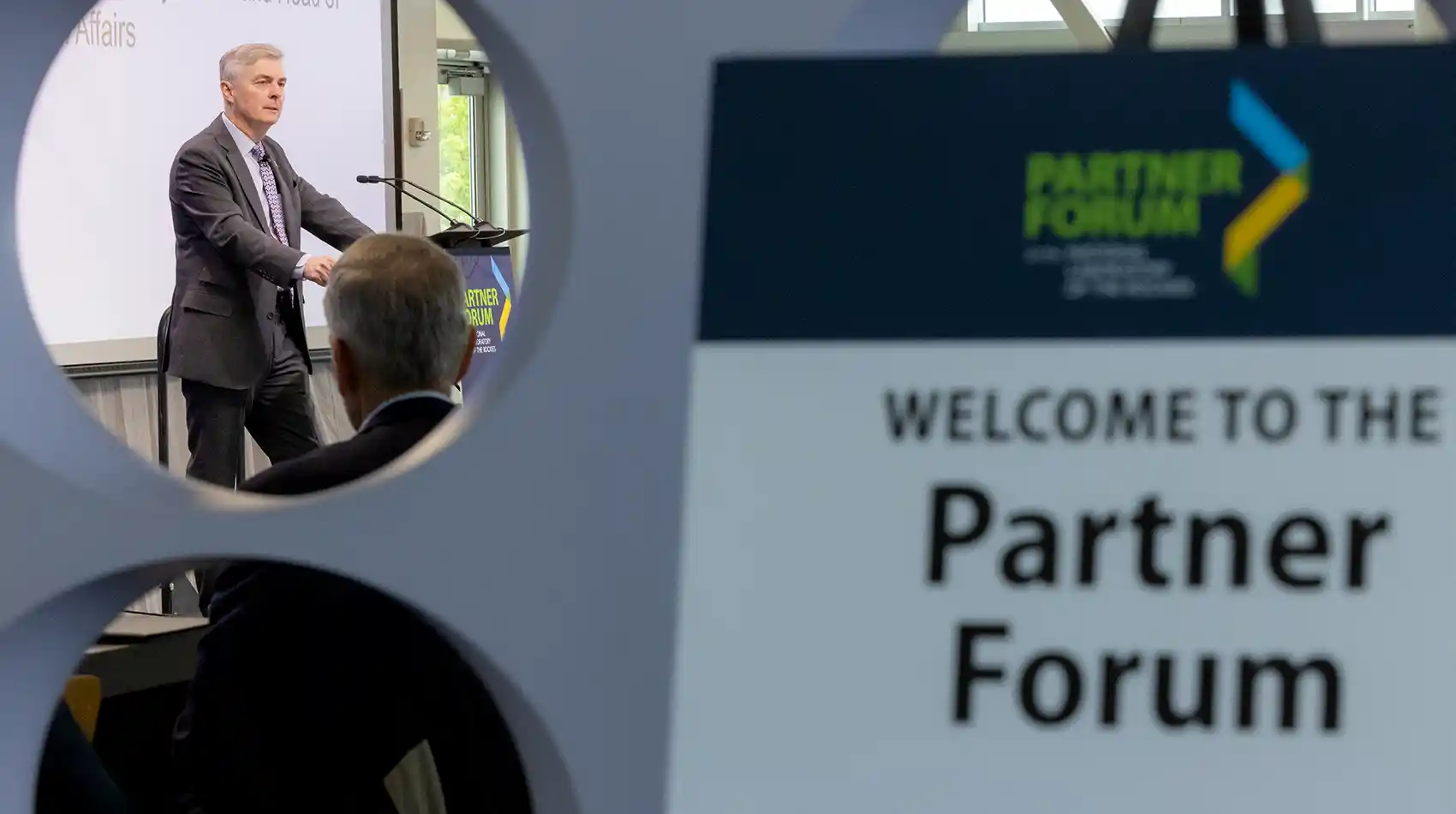 A person speaking at a podium with a sign with the text Welcome to the Partner Forum in the foreground.