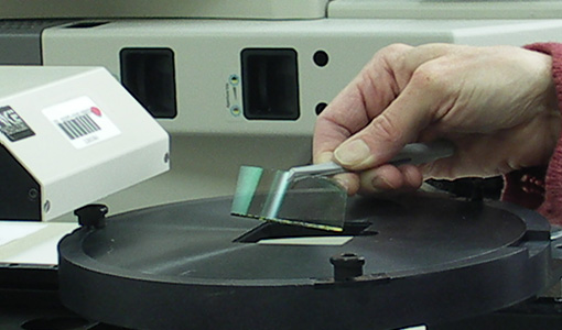 Hand placing a small film or coated sample onto a circular testing stage of a laboratory instrument.