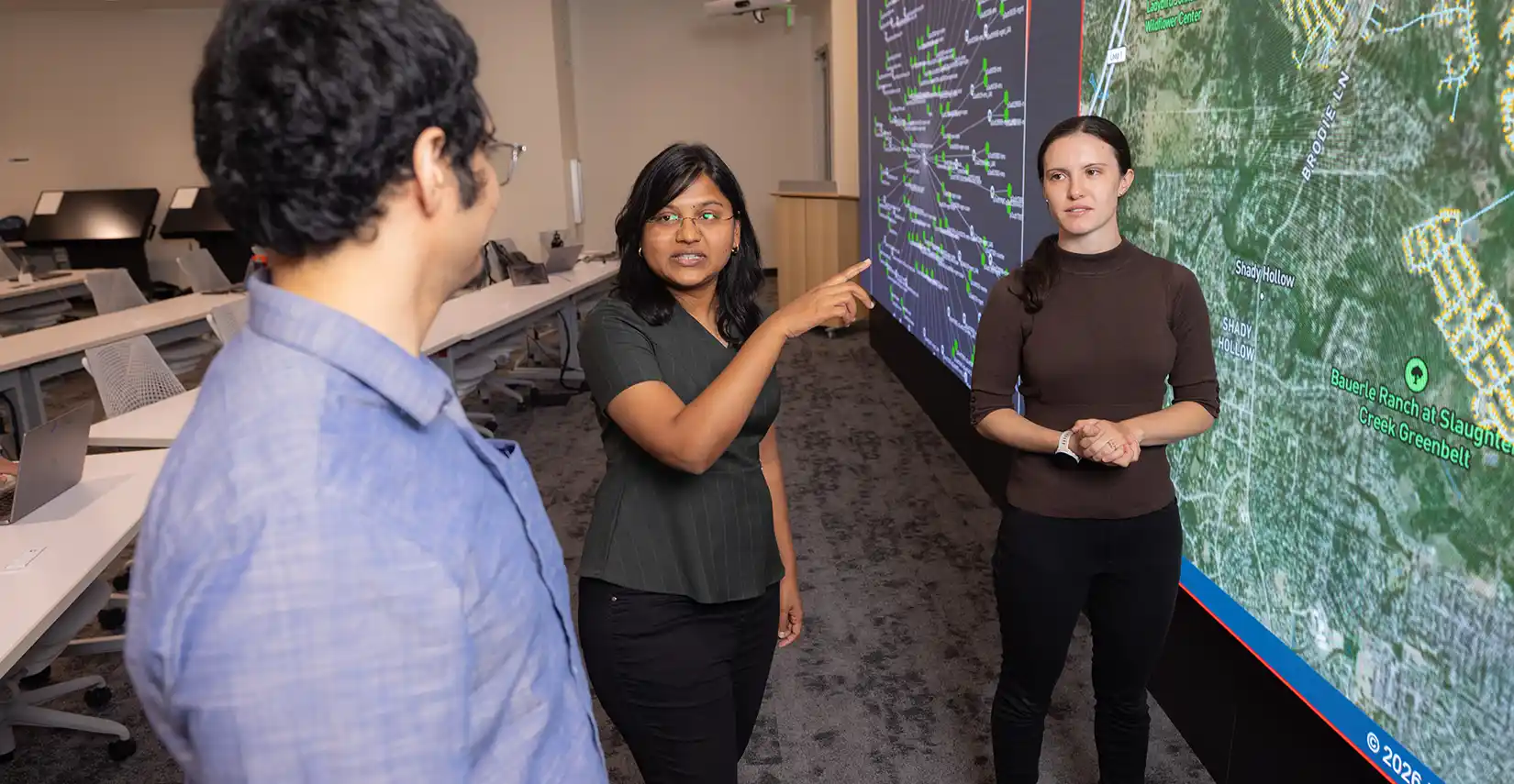 Three researchers look at large data screen, pointing and discussing map grid data.