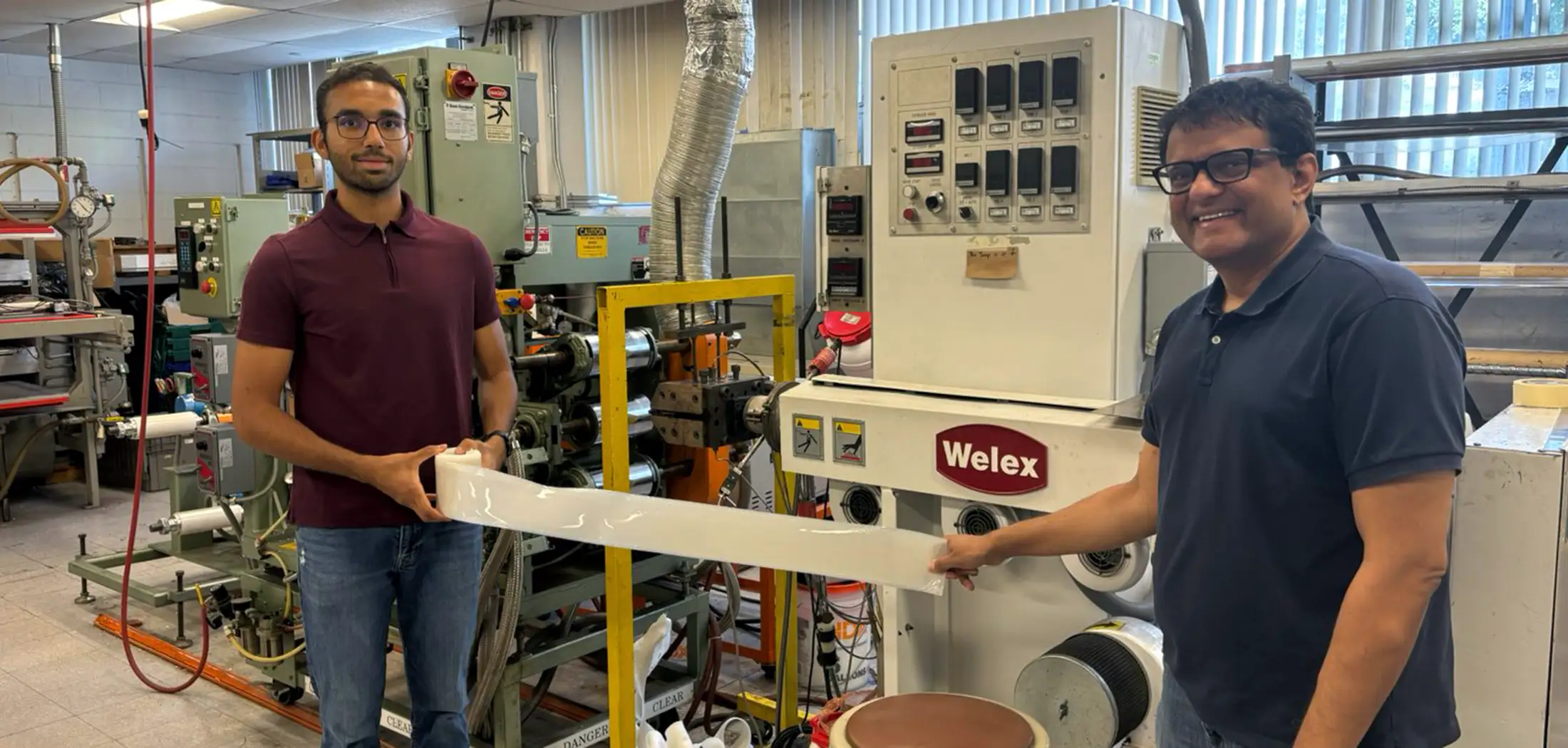 Two people stand in a lab setting by machine equipment holding white plastic and smiling at the camera.
