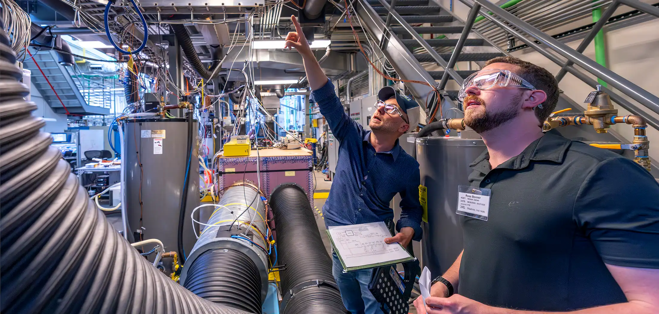 Two people stand in a laboratory surrounded by interconnected tubes, wires, and other equipment as one points upward while the other follows his gesture.