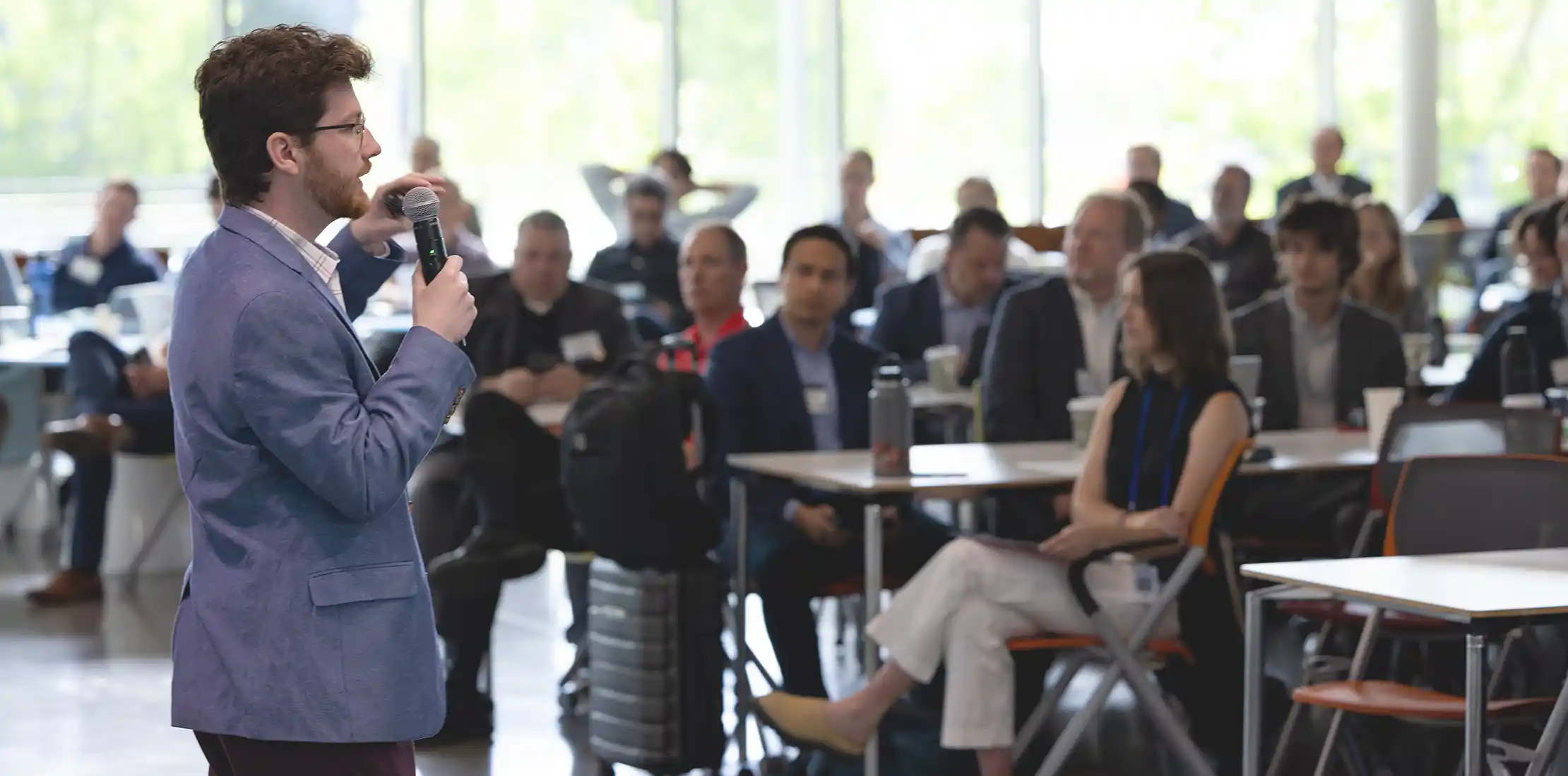 A man in business dress speaks into a microphone, addressing a room of people sitting at tables.
