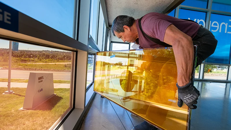 A person hunches down while sliding a large rectangular window into its slot in the bottom panel of a building wall.