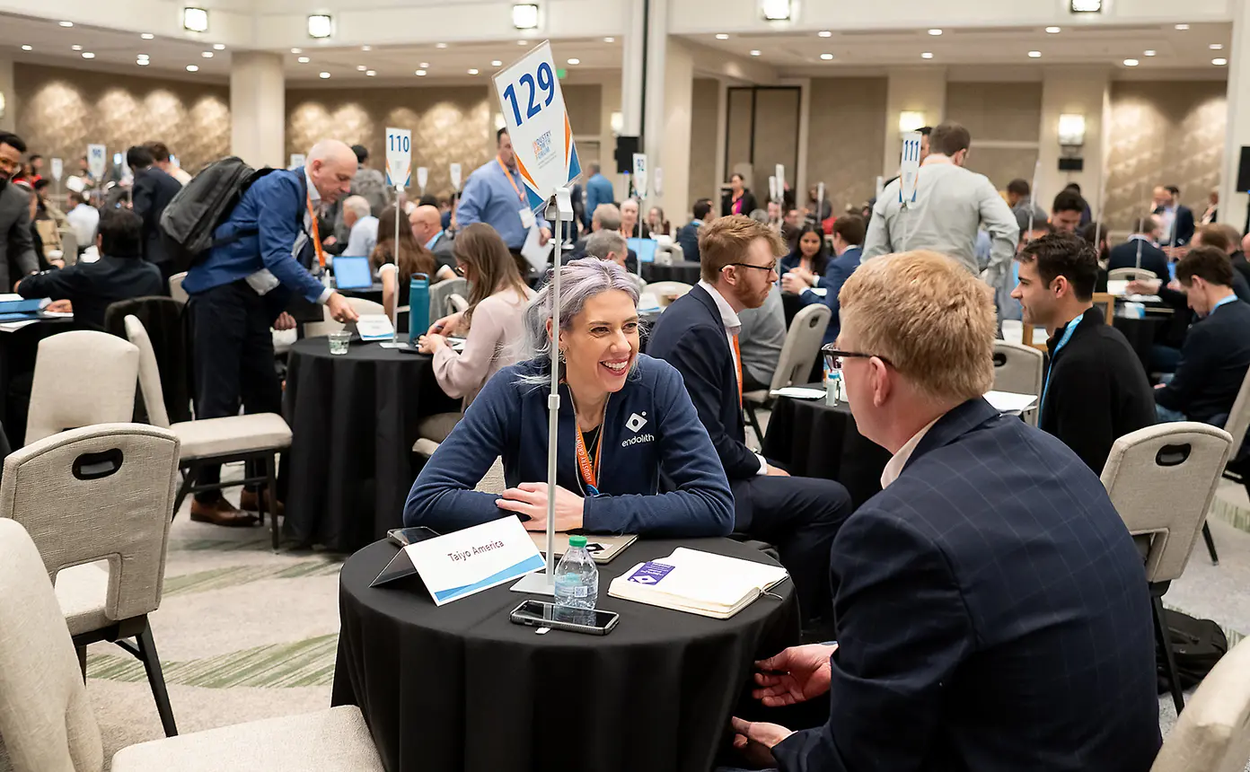 Two people, a CEO and an investor, sit across from each other at a small networking table, engaged in conversation, surrounded by a large, crowded room filled with hundreds of attendees having similar one-on-one discussions at mini tables.
