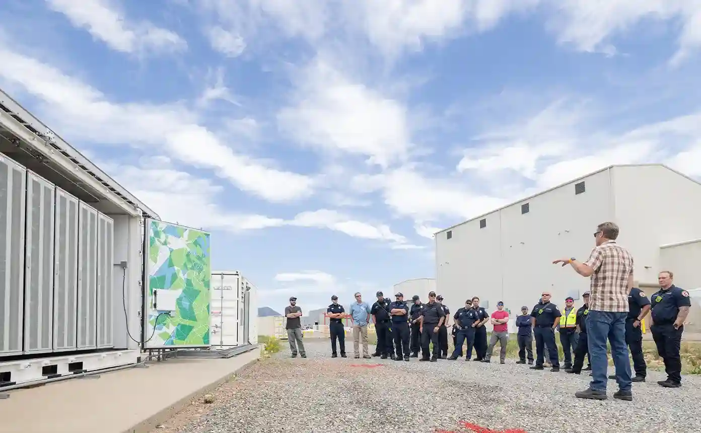 A large group of first responders stands next to a battery energy storage system.