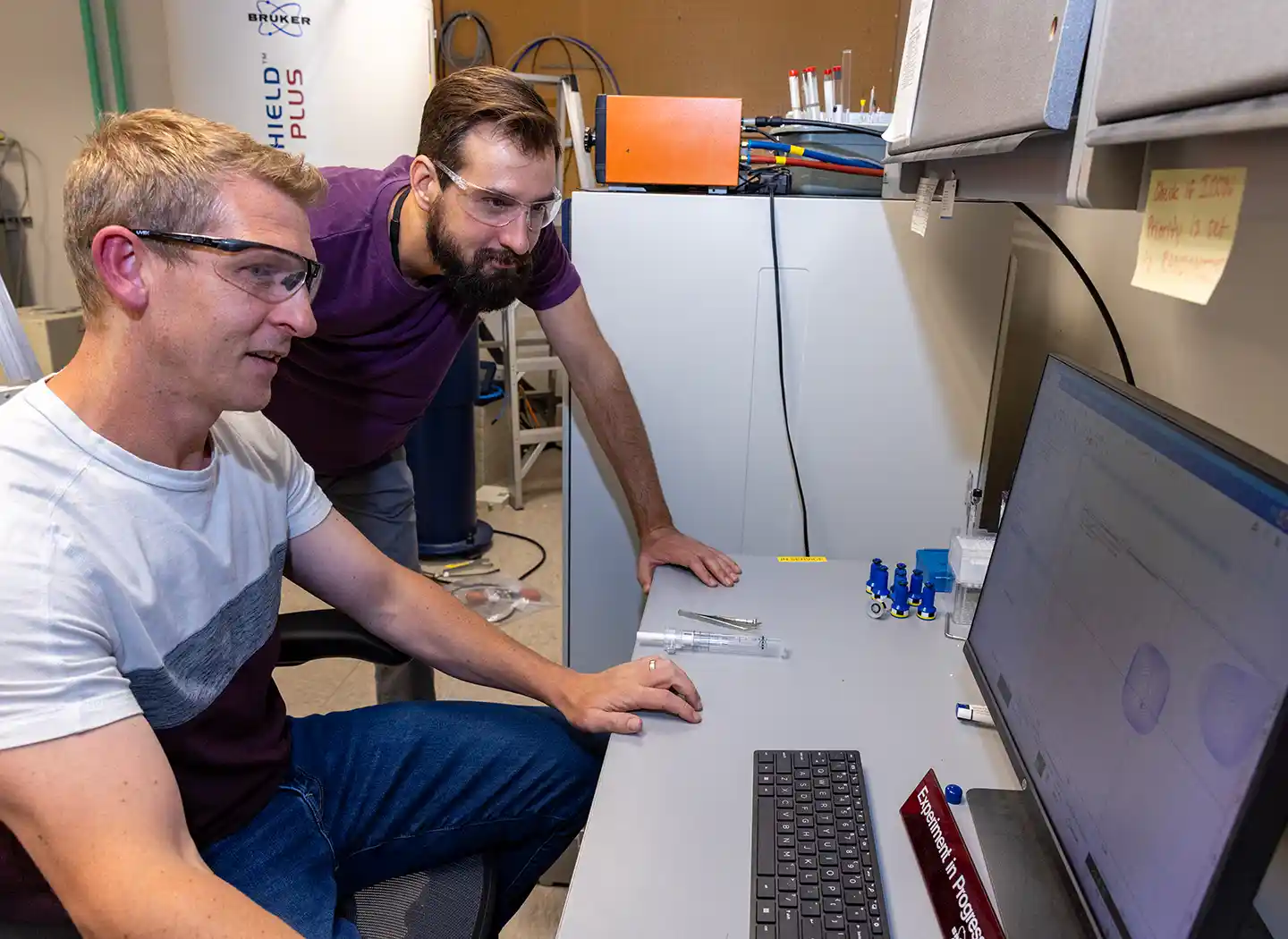 Two people in safety glasses look at a computer monitor.