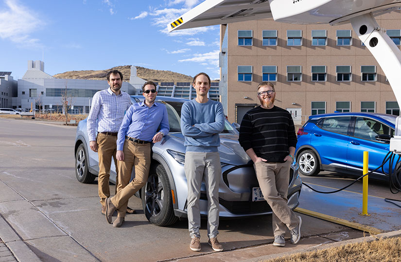 Four people stand in front of a charging car. 