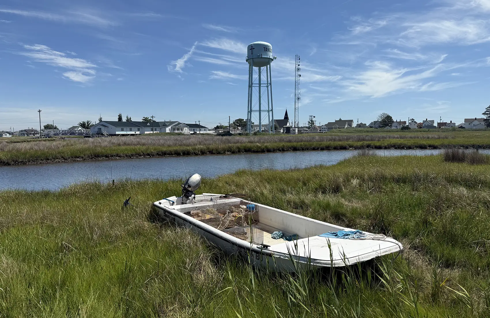 20260225-rising-above-the-tide-strategic-energy-planning-on-tangier-island-3221 A small motor boat with crabbing equipment sits on a grassy area in front of a waterway with a water tower behind it.