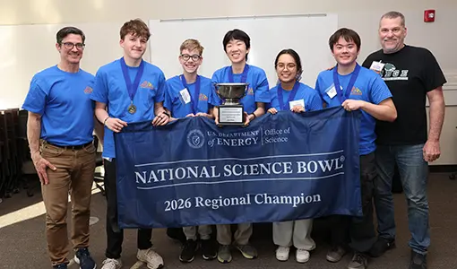 Continue reading about Seven people standing with a banner that reads "National Science Bowl: 2026 Regional Champion". Five of the people are student competitors wearing medals and the center student holds a trophy.