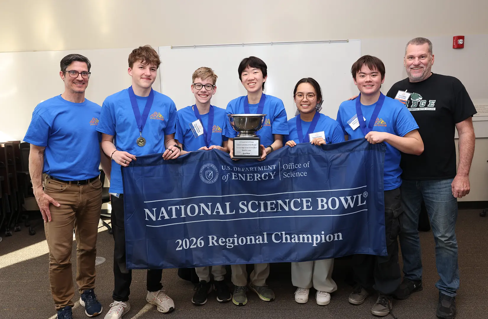 20260324-high-school-science-bowl-regional-champion-fossil-ridge-0A1A8414 Seven people standing with a banner that reads