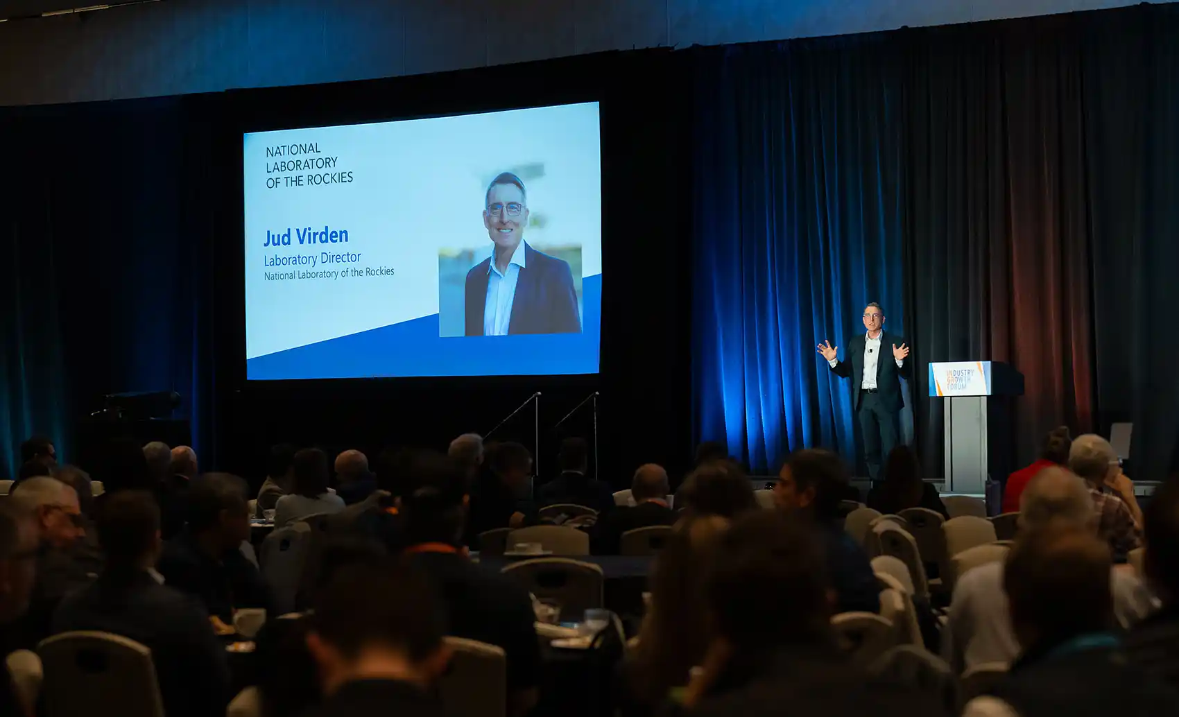 NLR director delivers a keynote presentation onstage in front of a curtain backdrop, with a large screen behind him displaying his photo and title.