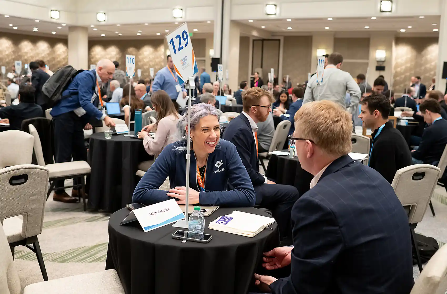 CEO and investor sit across from each other at a small networking table, engaged in conversation, surrounded by a large, crowded room filled with hundreds of attendees having similar one-on-one discussions at mini tables.