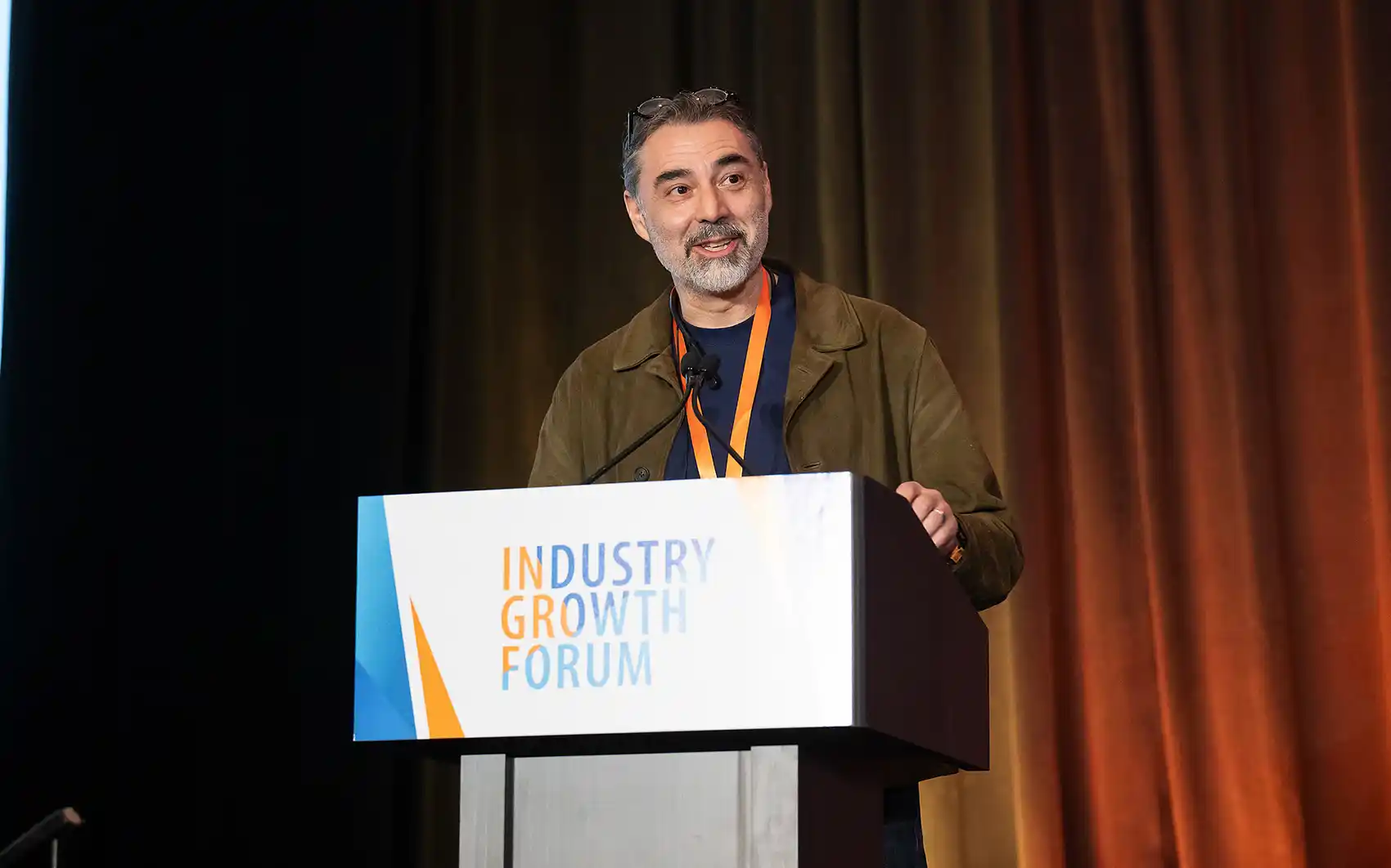 Man gives acceptance speech at IGF-branded podium with a curtain backdrop, wearing glasses, a brown jacket, and orange lanyard.