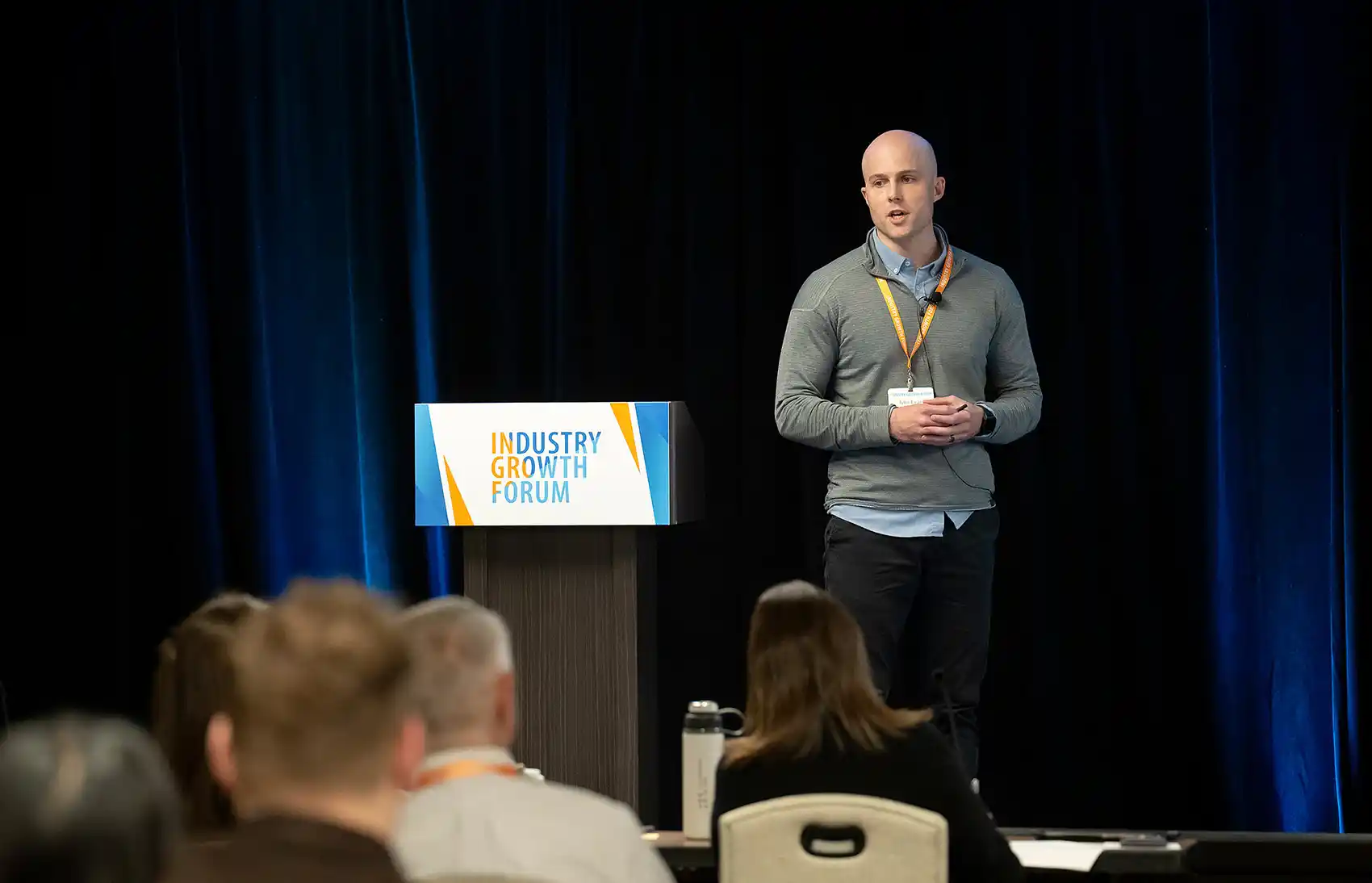 Against a blue-lit curtain backdrop, a man in a gray quarter-zip and orange lanyard delivers a pitch beside an IGF-branded podium.