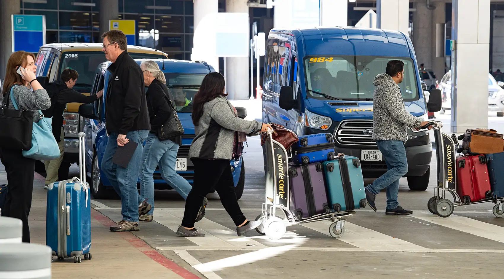 A busy airport arrivals intersection filled with people crossing a road carrying luggage and locating shuttles.