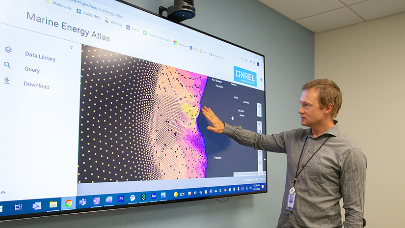 A man gesturing at a large screen that shows a visualization of wave energy resources off the Pacific coast