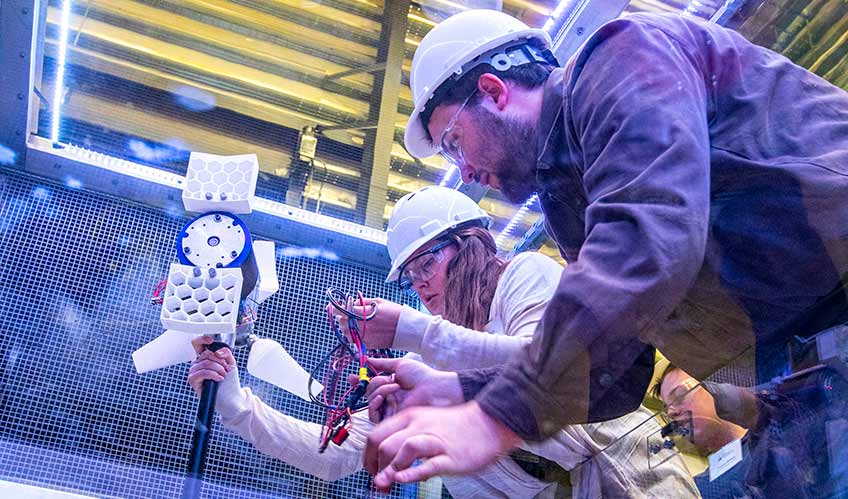 A young woman and young man in hardhats and safety glasses work on a small turbine inside a wind tunnel. 