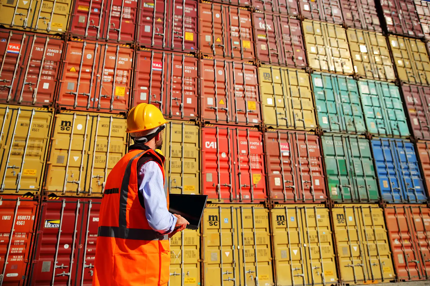 A man in a hard hat and safety vest inventories a tall, colorful stack of standardized metal shipping containers.