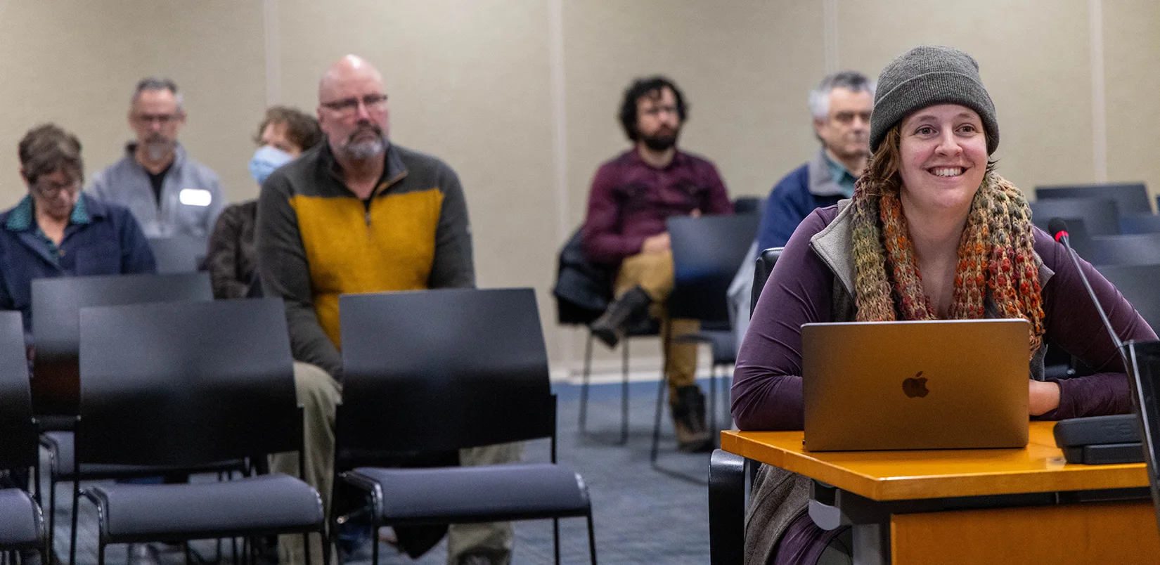 A woman sitting at a desk with a laptop and microphone to present while an audience behind her watches.