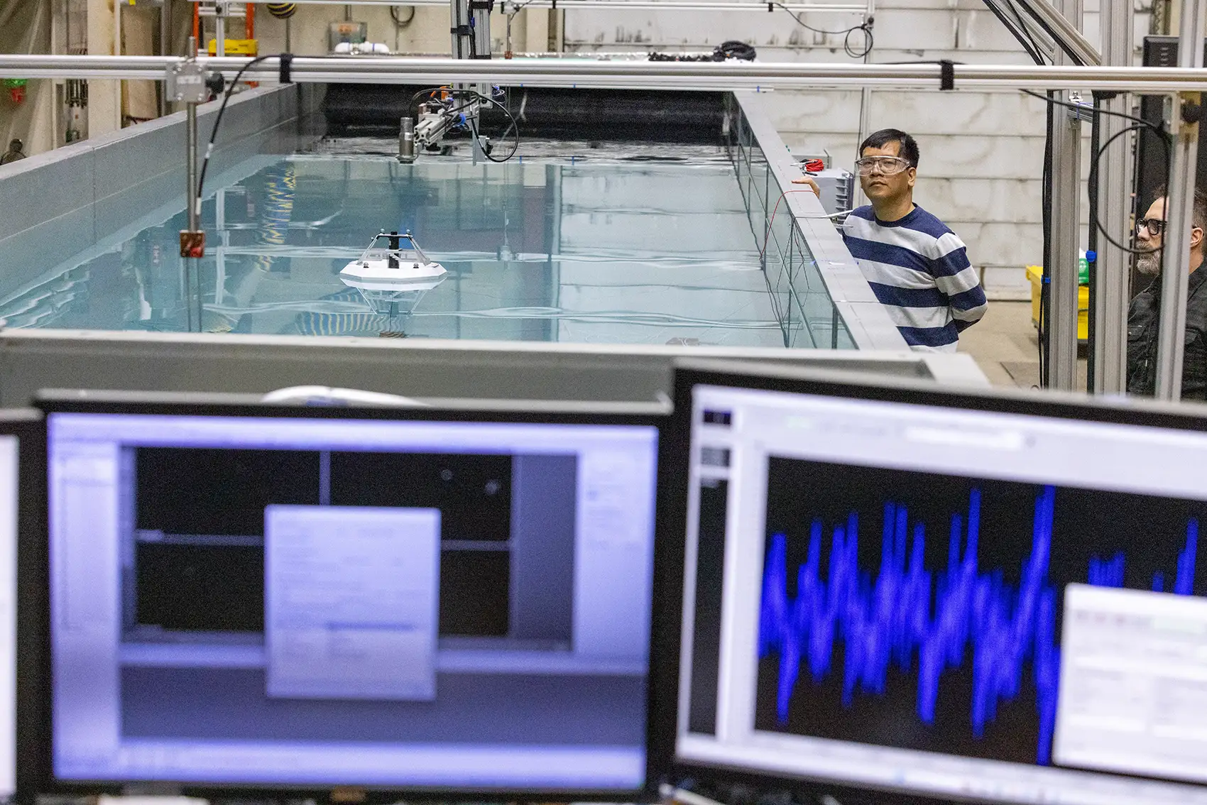 A person wearing safety goggles stands next to a large tank of water with a frisbee-sized device floating inside and computer screens visible in the foreground.