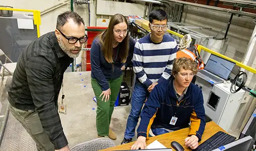 Continue reading about Four people stand or sit while looking at three computer monitors.