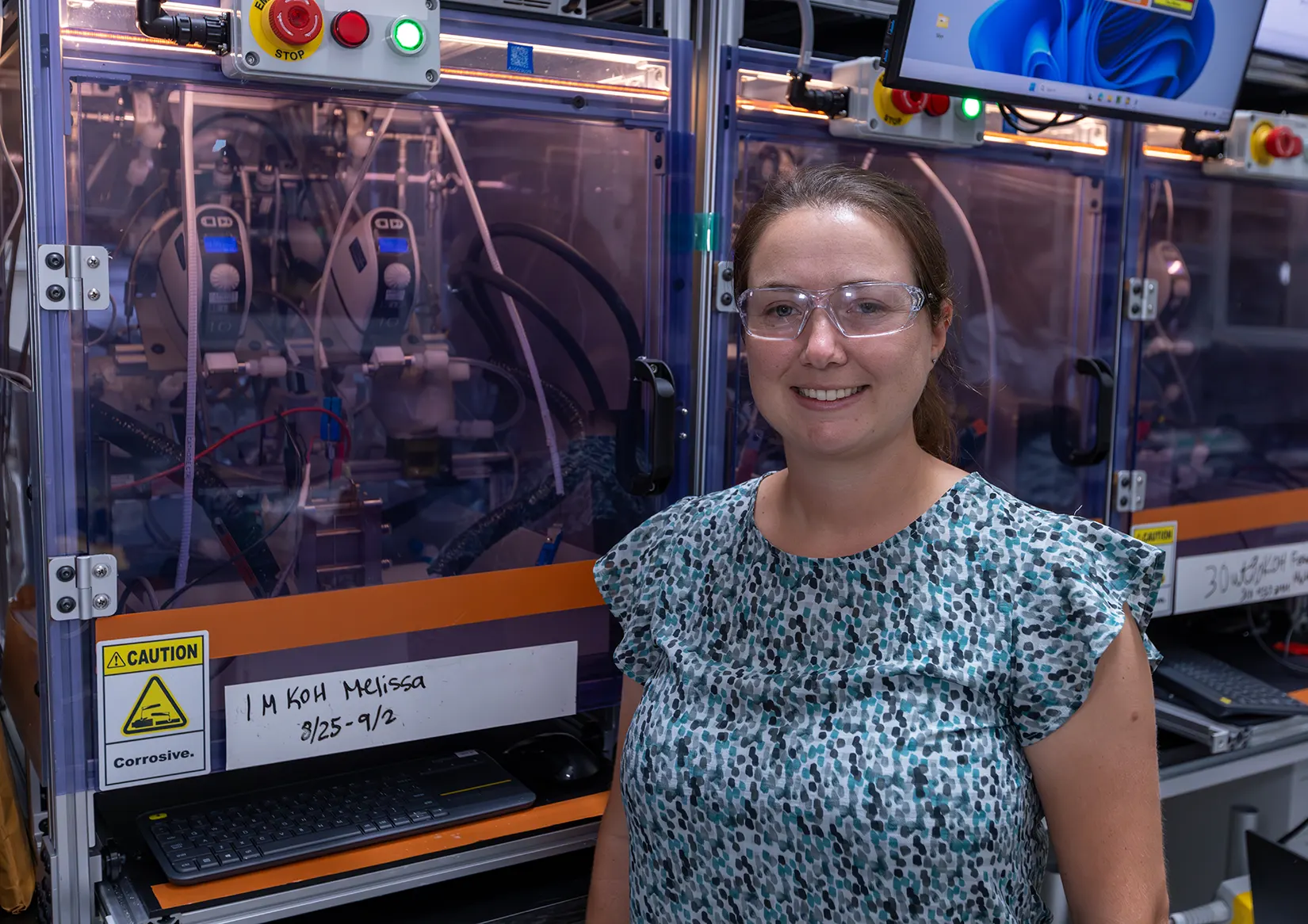 A woman with safety goggles stands in front of electrolyzer testing equipment.