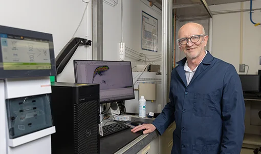 A man wearing a dark blue lab coat stands at a desk with a computer and machine with another screen.