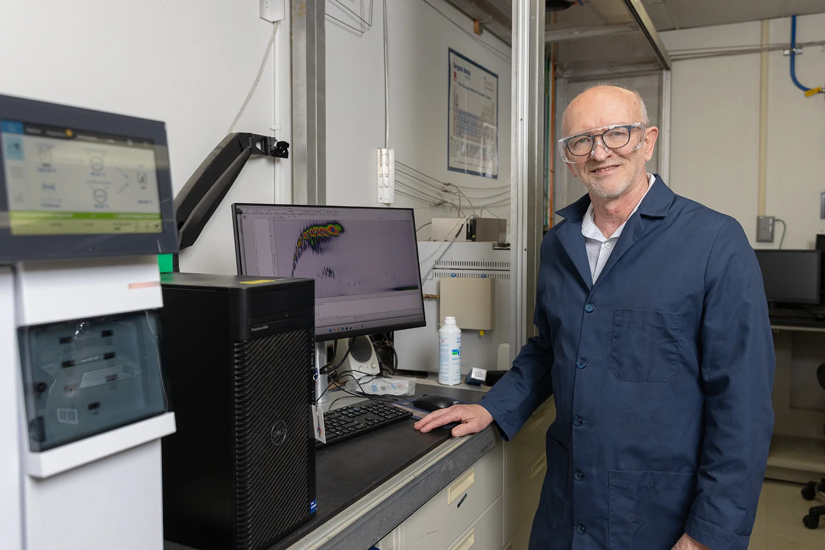 20260325-accomplished-researcher-recognized-by-major-industry-association-for-leadership-in-biofuels-research-robert-mccormick-tobi-104254 A man wearing a dark blue lab coat stands at a desk with a computer and machine with another screen.
