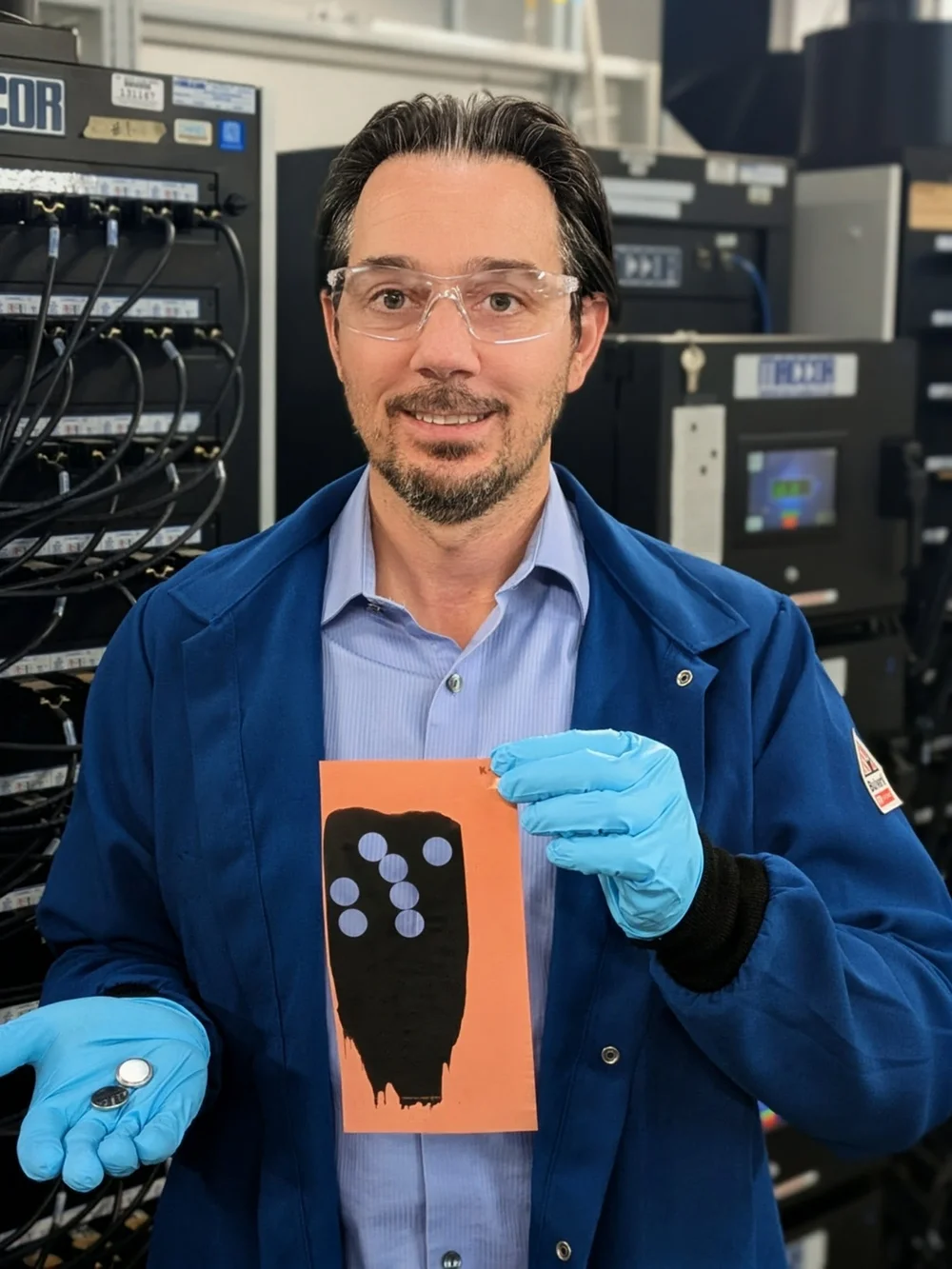 A researcher wearing blue gloves holds up a small graphite electrode and matching coin-sized batteries.