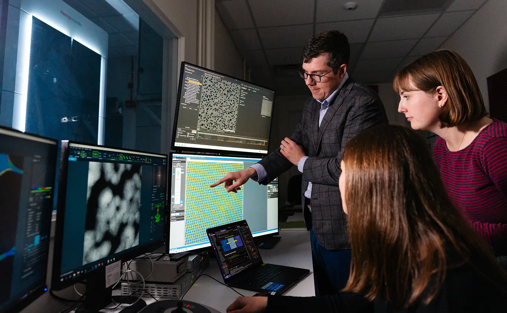 A man and woman standing and a woman seated, all looking at scientific information on multiple computer monitors.