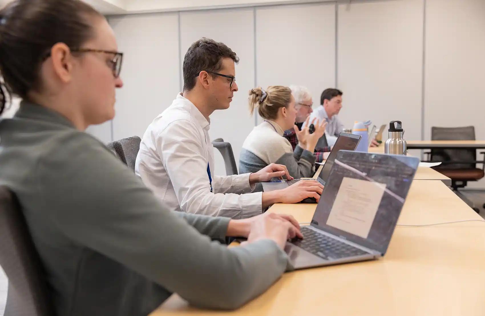 Several people sit at a desk with their laptops open.