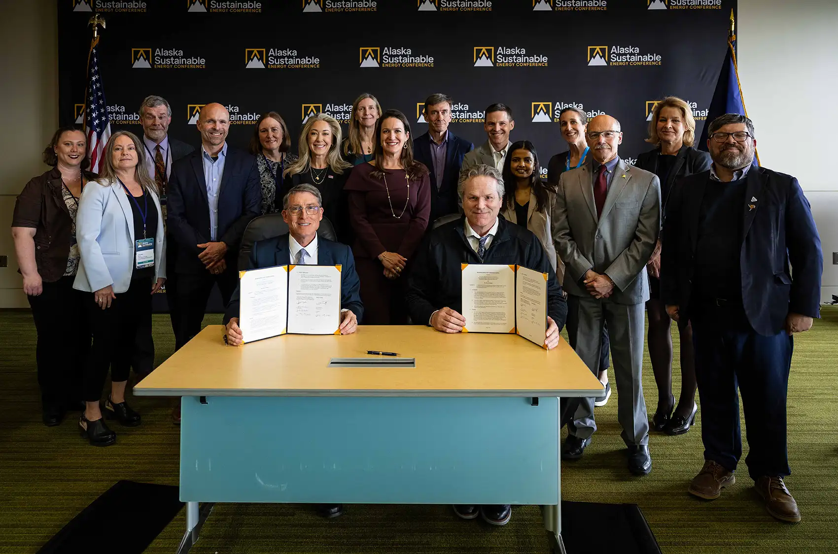20260630-new-partnerships-with-state-of-alaska-and-university-of-alaska-fairbanks-expand-on-critical-minerals-and-energy-innovation-1431 Two people sit at a desk, holding up folders with paper. A large group of people stand behind them.