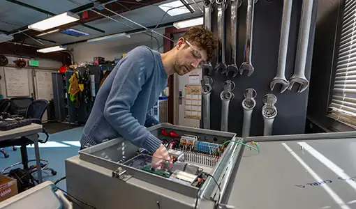 Continue reading about A young man wearing safety glasses and a blue sweater leans over a large, open gray electrical control box on a workbench.