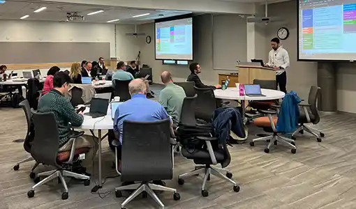 Continue reading about A group of people sitting around tables in a room while a person stands at a podium giving a presentation.
