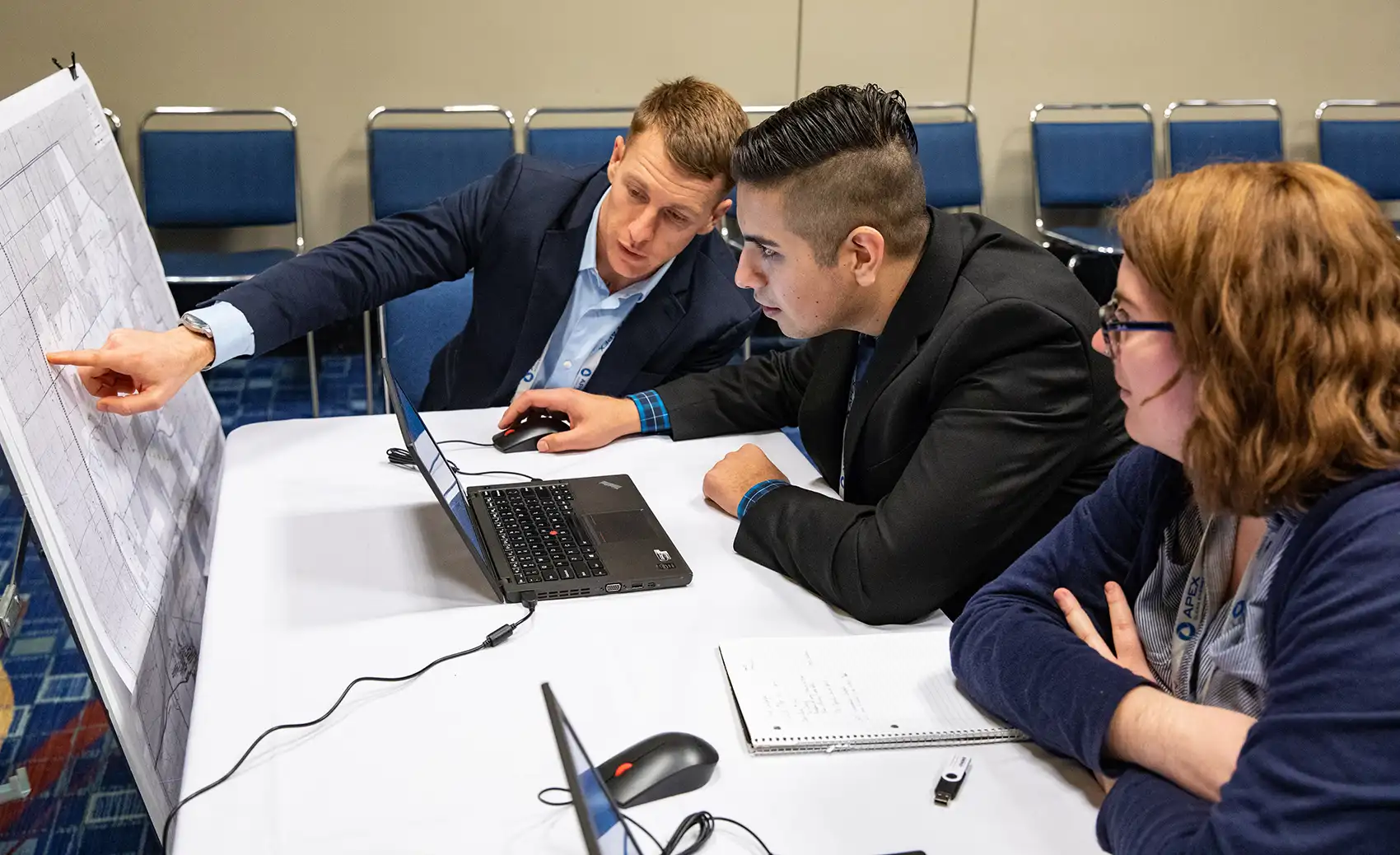 Three people seated at a table with laptops, a notepad, and a thumb drive study information printed on a table-length section of poster board propped up in front of their table.