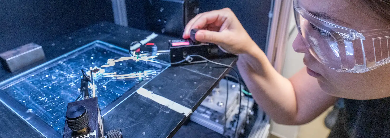 Researcher in safety goggles operating a probe station over an illuminated microfluidic chip in a laboratory.