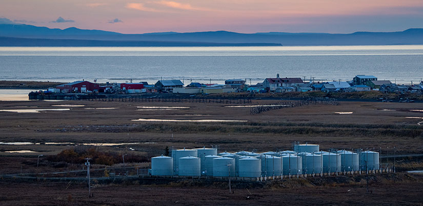 alaska-community-energy-storage Buildings and homes line a waterfront with tanks in the foreground and mountains in the background