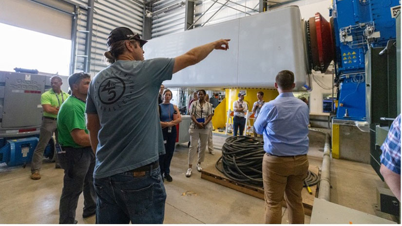 20240731-newsletter-hydrovision A person in a hat points to a large wind turbine gearbox attached to a dynamometer while a tour group looks on