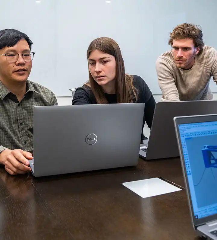 Three people look at a laptop around a conference table.