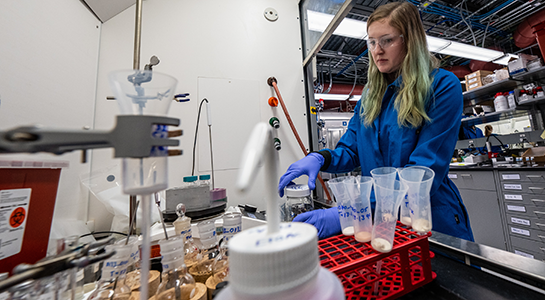 A person wearing protective eyewear, gloves, and a lab coat handles materials within a laboratory.